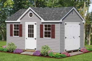 Dormer style shed with gray vinyl siding, a white front door with window panes, 2 windows with red shutters, gray roofing, and white double doors on the shed's right side.