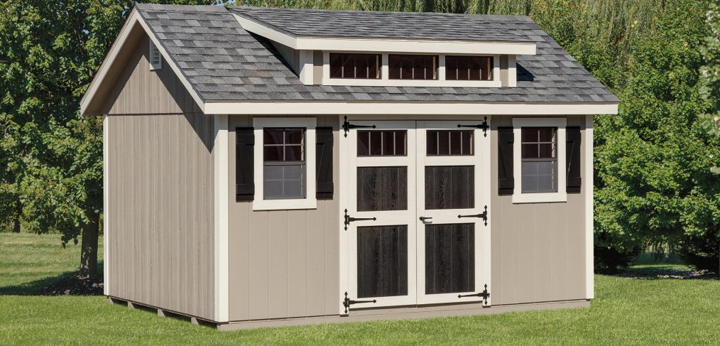 Transom dormer shed with tan siding, white trim, black double doors, 2 windows with black shutters, gray roofing, and transom dormer windows.