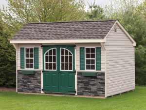 Quaker style shed with beige vinyl siding, green double doors with curved window panels, 2 windows with green shutters and green window boxes, brown roofing, and stone accents on the front of the building.