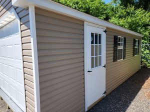 Garage with tan siding, white trim, a white panel garage door, a white side door with windows and a lock, and white windows with black shutters.