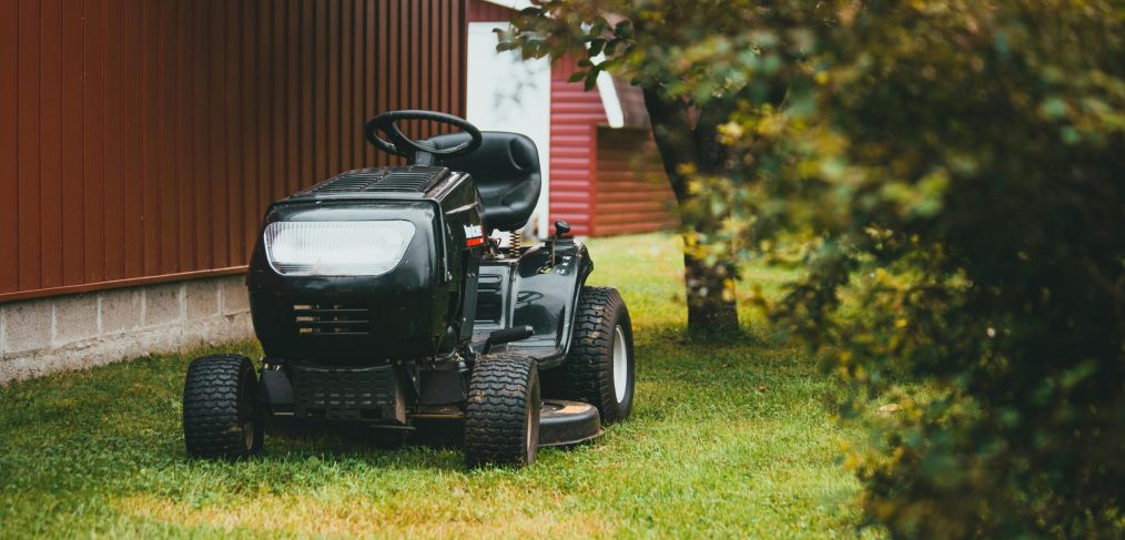 Black riding mower sitting in grass next to a red building with a matching red and white shed in the background.